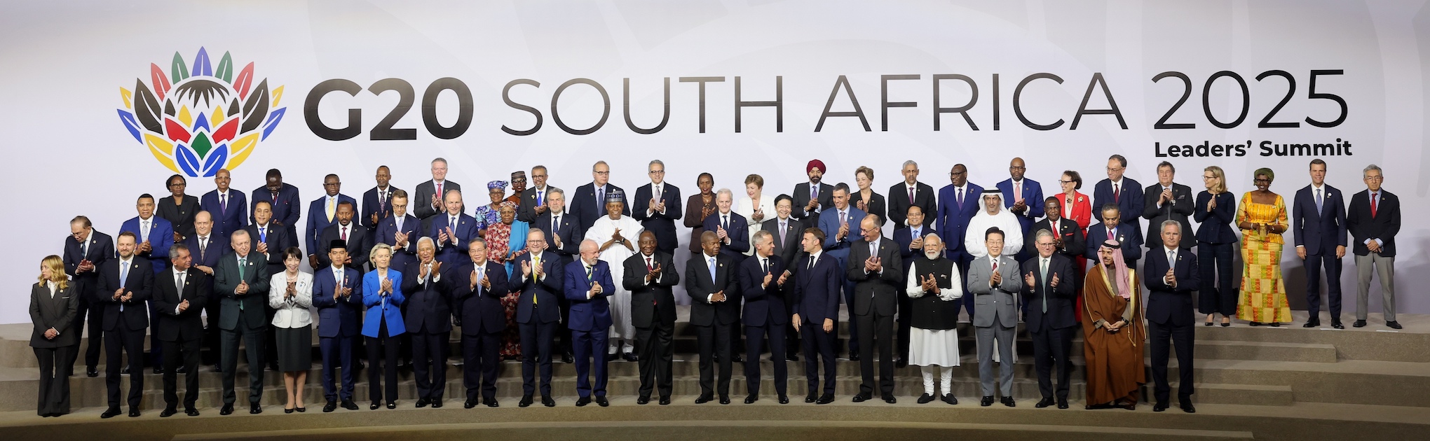 Official group photograph of G20 heads of state, government, and delegated representatives at the 2025 Johannesburg Summit. Approximately 25 leaders, predominantly men in dark suits, stand and sit in three rows on a stage, smiling and applauding. Behind them is a massive blue backdrop with the G20 South Africa 2025 logo and the words “Solidarity, Equality, Sustainability.” Many of the nations represented here – including the United States, Russia, China, France, and Germany – are the world’s largest arms exporters and are directly or indirectly involved in prolonging the wars in Gaza, Ukraine, Sudan, and the Democratic Republic of Congo, conflicts that together received only four sentences in the summit’s final 122-point declaration.