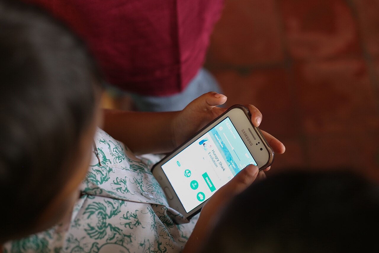 A young Indonesian boy sitting outdoors focused on a smartphone while downloading a game, representing children’s heavy mobile and social media use in Indonesia.
