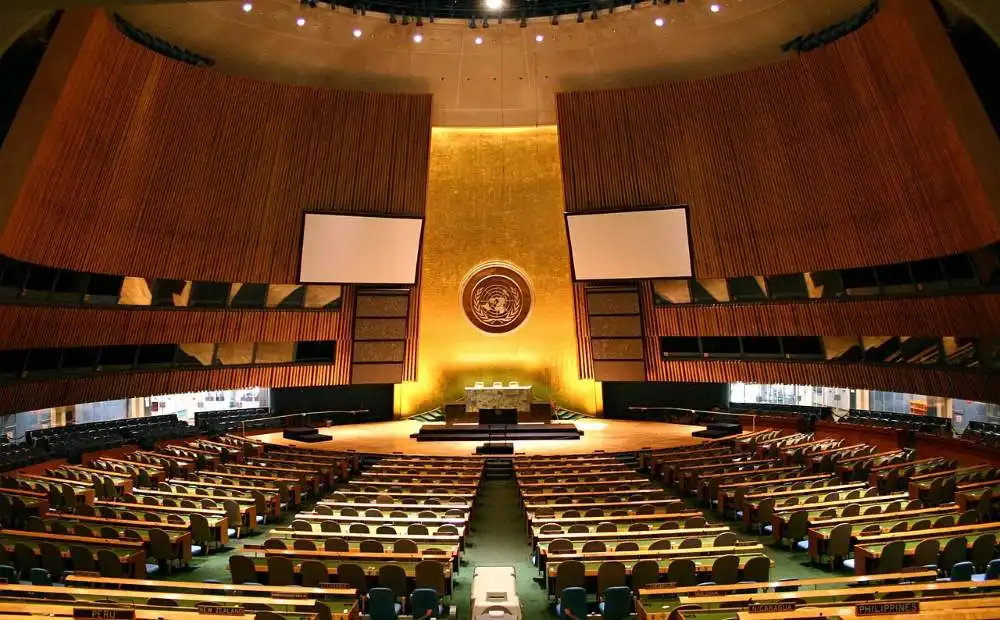 Empty United Nations General Assembly hall in New York, with rows of seats and the iconic UN emblem at the front.