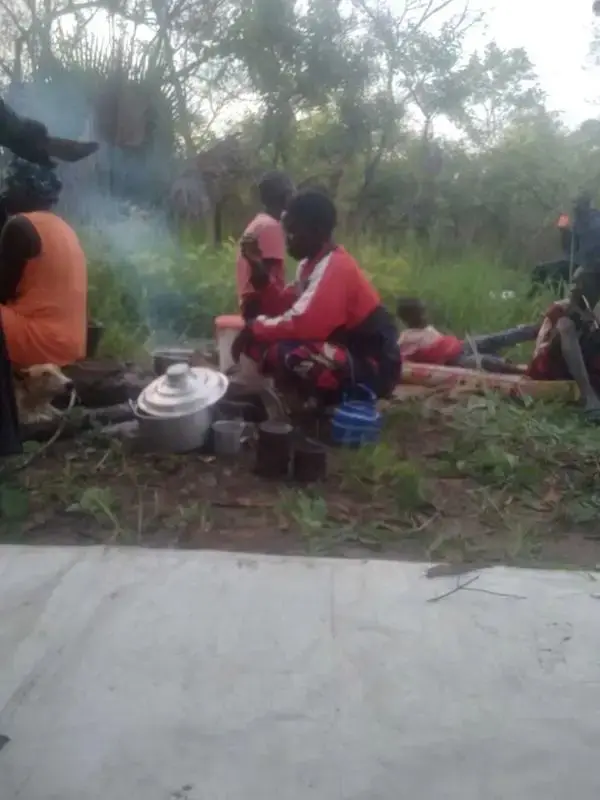 A South Sudanese displaced family cooking a meal over an open wood fire in the bush after fleeing armed violence in 2026, surrounded by minimal possessions in a makeshift outdoor shelter.